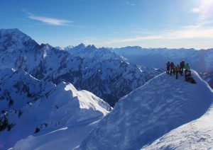 Cordillera de montañas nevadas. Un grupo de personas las divisa desde la cima de una de las montañas.